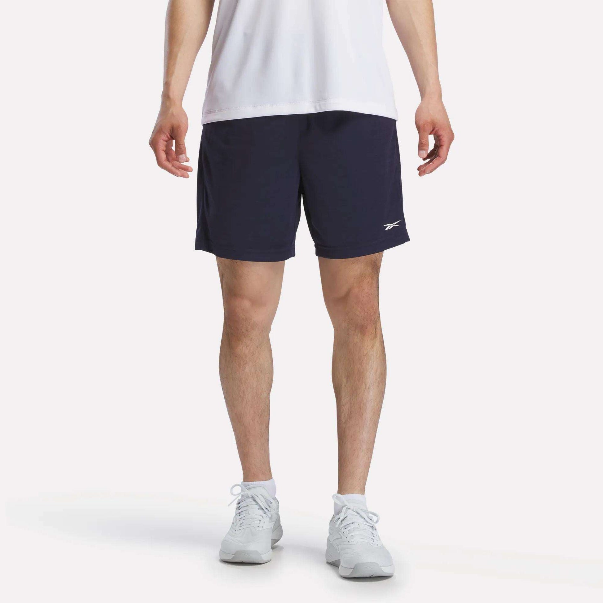 A person wearing a white T-shirt, white socks, and white sneakers is standing against a plain white background, paired with Reebok Men's Inclination Shorts in navy blue featuring a small logo.