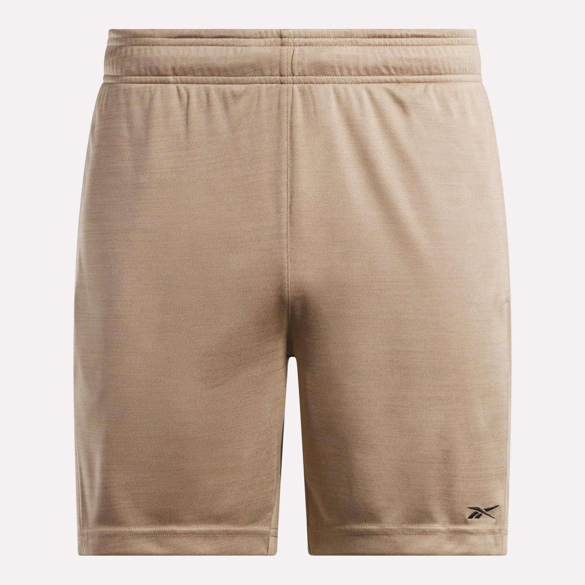 The Reebok Men's Inclination Shorts feature tan, soft fabric and an elastic waistband. A small black Reebok logo accents the lower left leg. These athletic shorts are shown against a plain, light background.