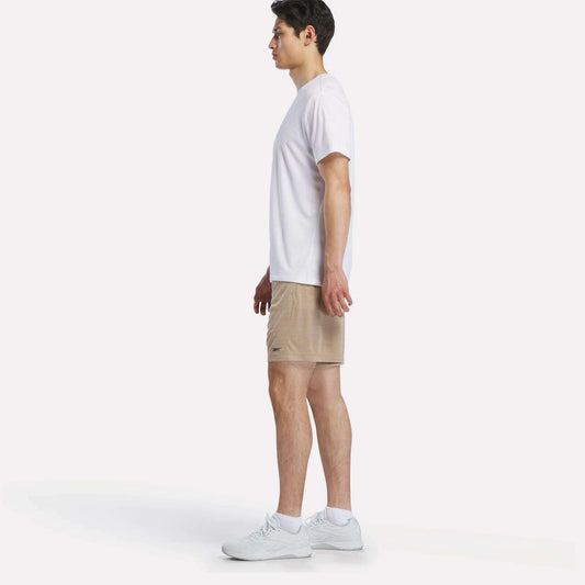 A man stands in profile against a white background, wearing a white T-shirt, Reebok Men's Inclination Shorts, and white sneakers.