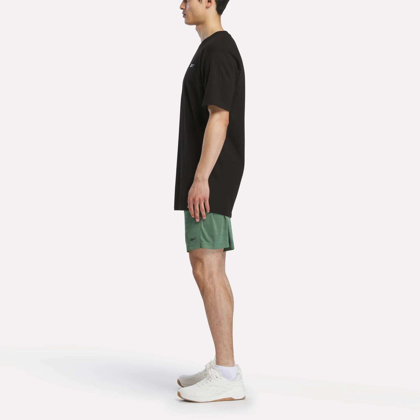 A person stands in profile against a plain white background, wearing a black t-shirt, Reebok Men's Inclination Shorts in green, and white athletic shoes.