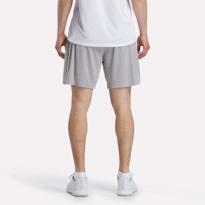 A person is shown from the waist down against a plain light background, wearing a white shirt, Reebok Men's Inclination Shorts in light gray, and white sneakers.