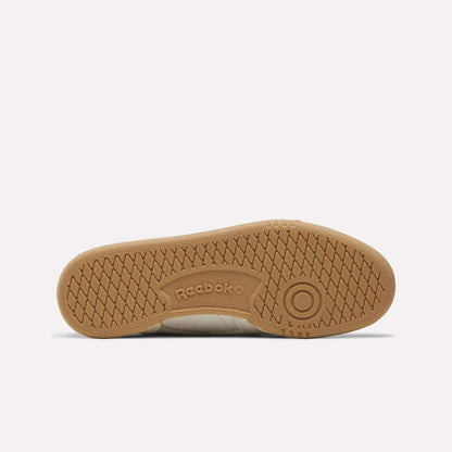 The image shows the sole of the Reebok Women's Club C Revenge Shoes, highlighting a light brown gum rubber outsole with a herringbone tread pattern and the Reebok logo embossed in the center.