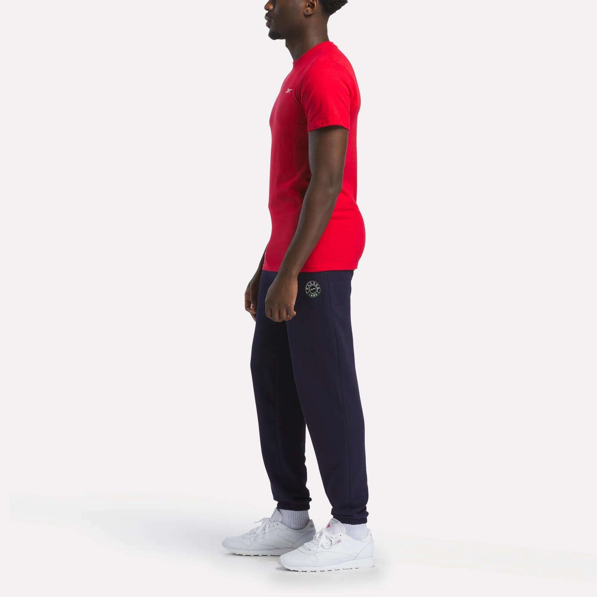 A person stands in profile against a white background, wearing a relaxed fit Reebok Men's Small Logo T-Shirt, navy jogger pants with a small logo on the hip, and white sneakers.