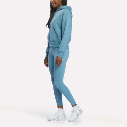 A woman faces left in a Reebok Women's Cropped Full-Zip Hoodie, styled with matching blue leggings and white sneakers, set against a plain white background.