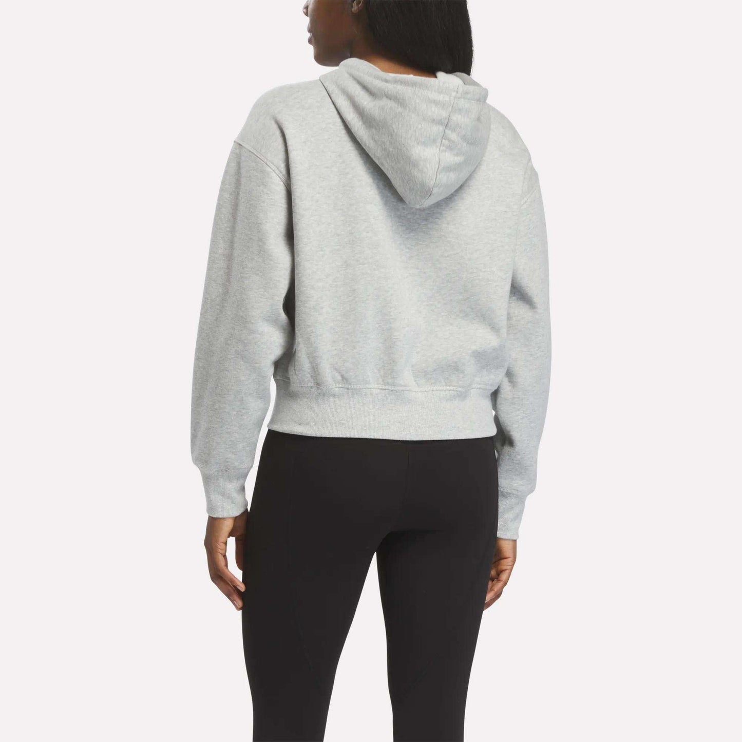 A woman is seen from the back, wearing a light gray Reebok Women's Cropped Full-Zip Hoodie and black leggings, standing against a plain white background.