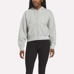 A person wears a Reebok Women's Cropped Full-Zip Hoodie in Grey Heather, featuring front pockets, paired with black leggings against a plain light background.