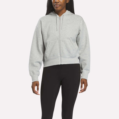 A person wears a Reebok Women's Cropped Full-Zip Hoodie in Grey Heather, featuring front pockets, paired with black leggings against a plain light background.