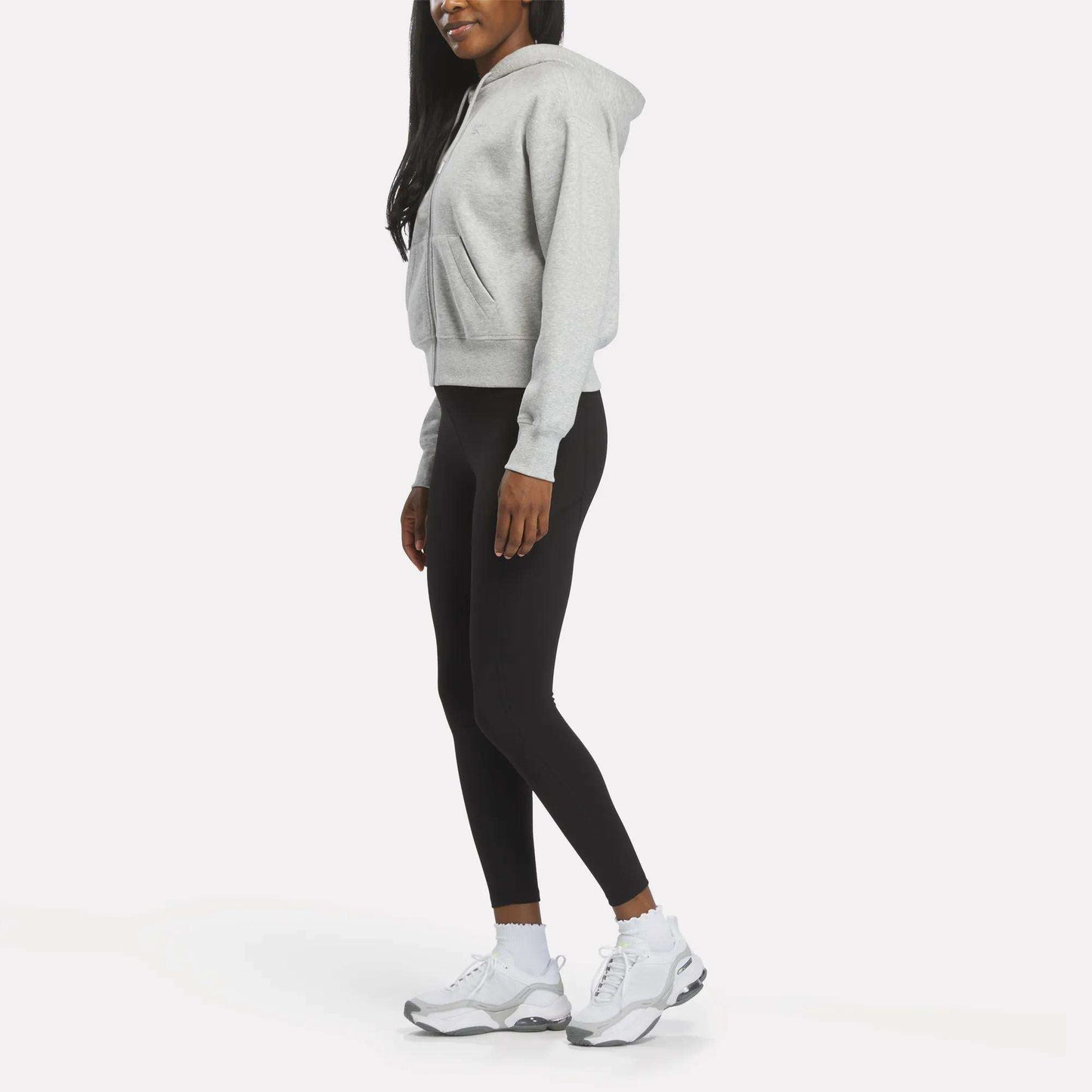 A woman models the Reebok Women's Cropped Full-Zip Hoodie in gray, paired with black leggings and white sneakers, standing sideways against a white background with her hands relaxed at her sides and her face partially out of frame.