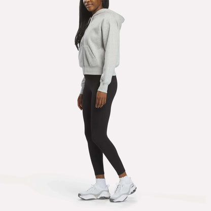 A woman models the Reebok Women's Cropped Full-Zip Hoodie in gray, paired with black leggings and white sneakers, standing sideways against a white background with her hands relaxed at her sides and her face partially out of frame.