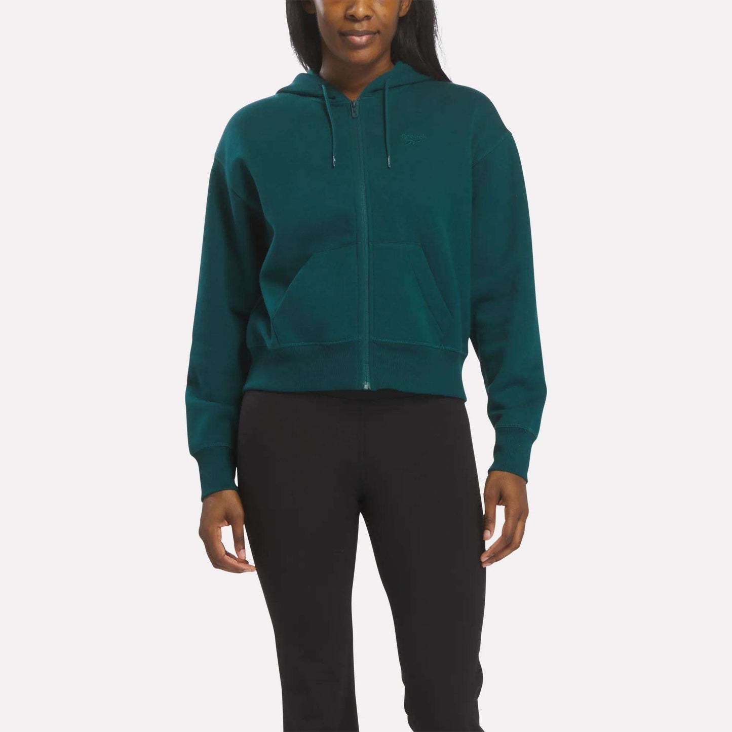A woman faces forward in a metal green Reebok Women's Cropped Full-Zip Hoodie with a hood and front pockets, paired with black pants, against a plain white background.