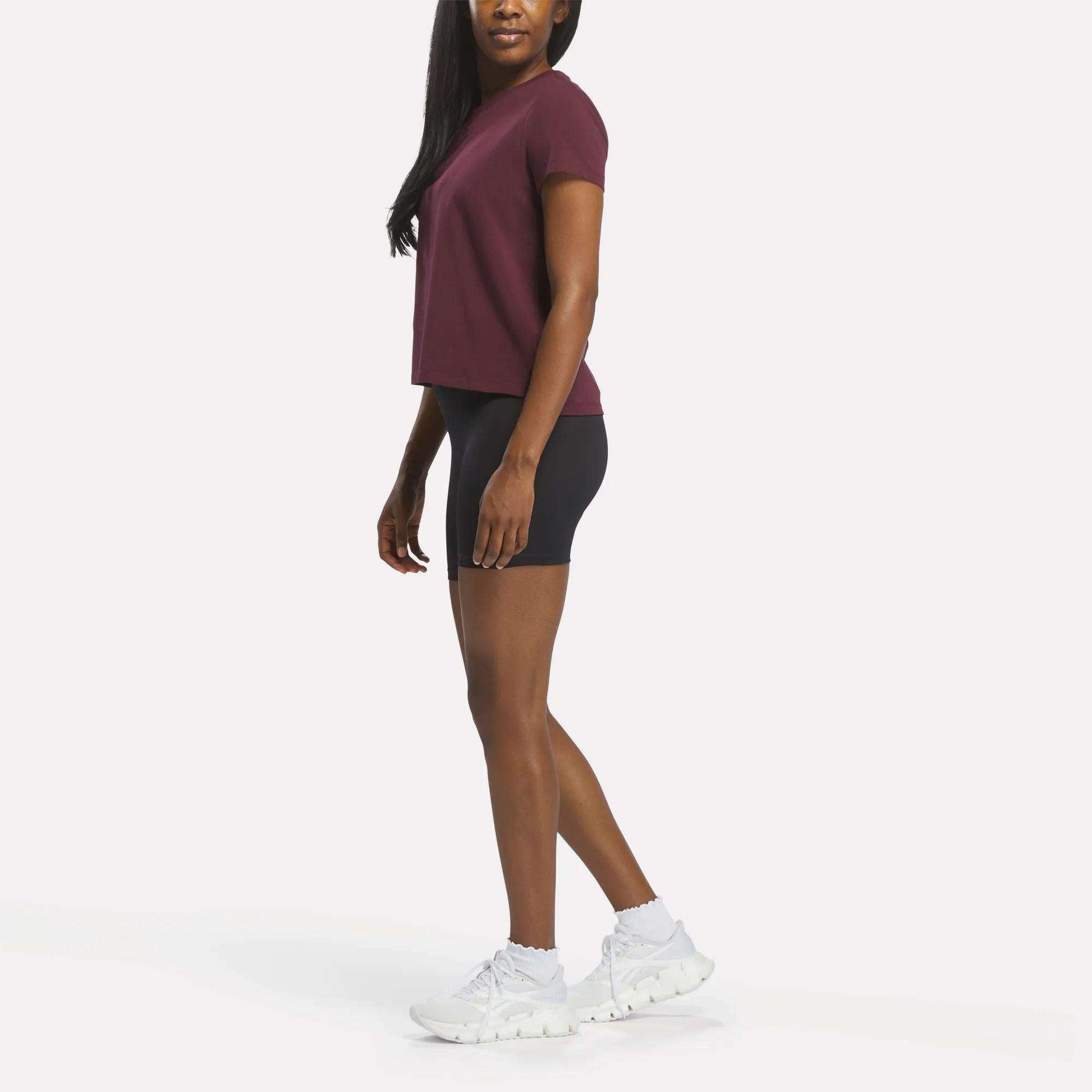 A woman stands sideways, wearing the Reebok Women's Graphic Glow Top in maroon, black fitted shorts, and white athletic shoes against a plain white background.