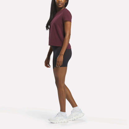 A woman stands sideways, wearing the Reebok Women's Graphic Glow Top in maroon, black fitted shorts, and white athletic shoes against a plain white background.