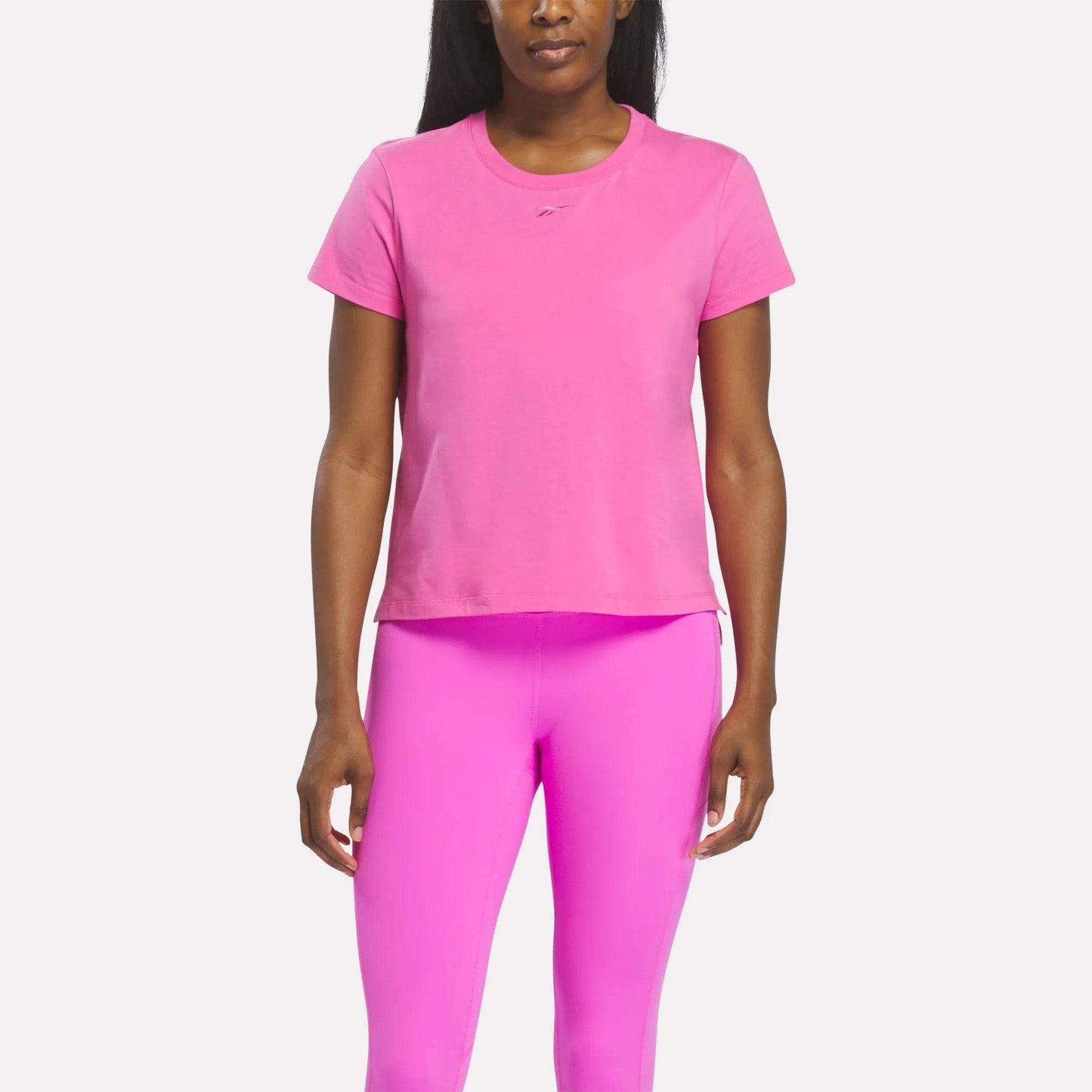 A person stands facing forward wearing a Vector Virtual Pink Women's Graphic Glow Top by Reebok with matching pink leggings against a plain white background.