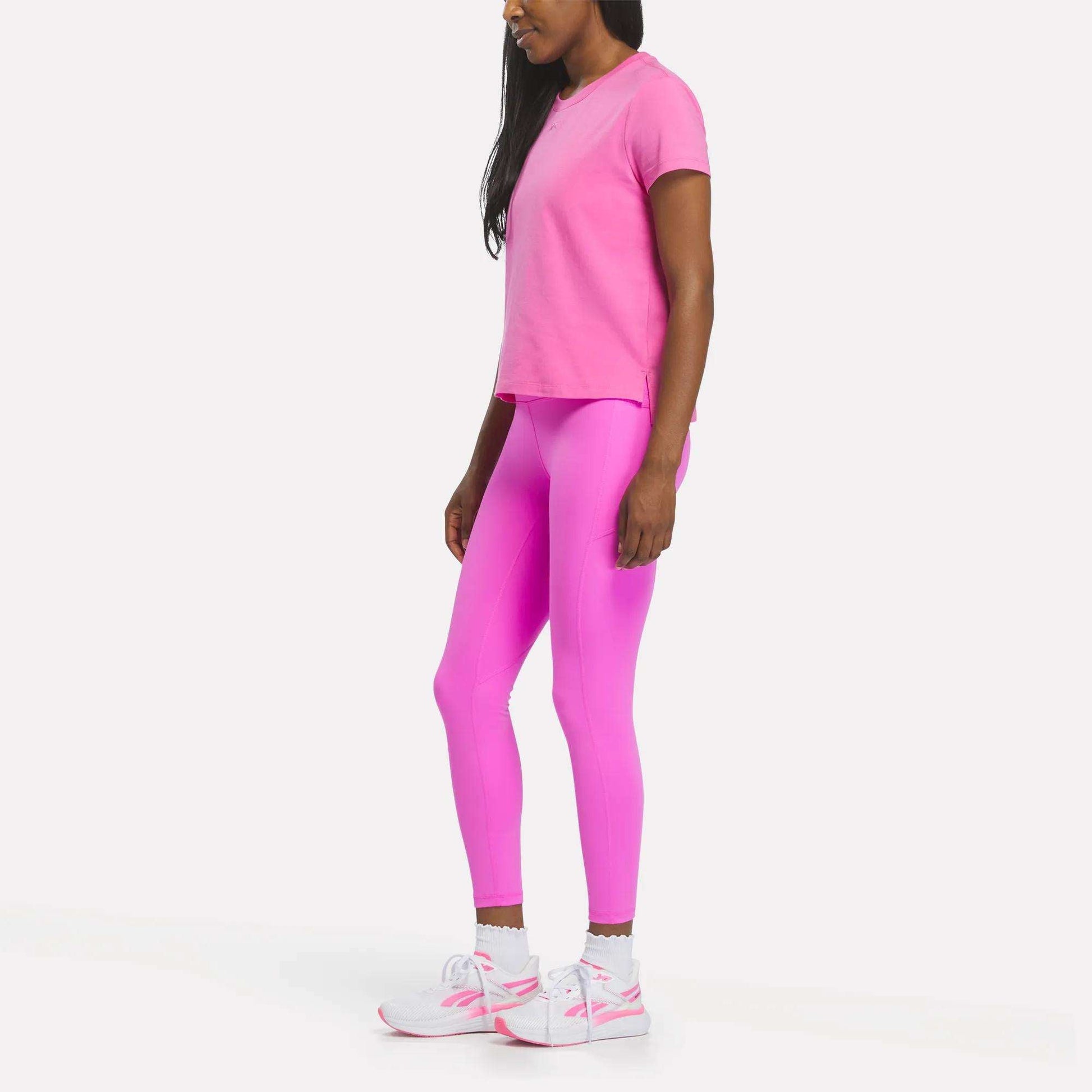 A woman stands sideways against a plain white background, wearing a bright pink Reebok Women's Graphic Glow Top, matching pink leggings, and white sneakers with pink accents.