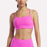 A woman models the Reebok Women's Low-Impact Sports Bra with adjustable straps in bright pink, paired with matching high-waisted leggings, against a plain white background. The image crops from her shoulders to just above her knees.