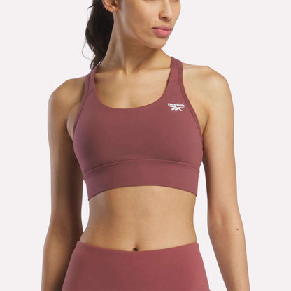 A woman in a mauve Reebok Women's Rush Medium-Impact Sports Bra and matching high-waisted leggings poses against a plain, light background, showcasing activewear suited for medium-impact workouts.