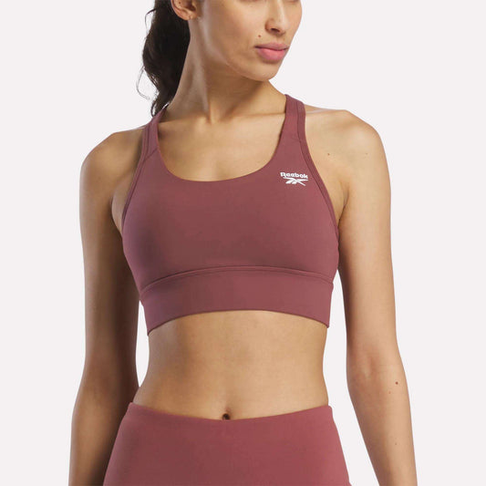 A woman in a mauve Reebok Women's Rush Medium-Impact Sports Bra and matching high-waisted leggings poses against a plain, light background, showcasing activewear suited for medium-impact workouts.