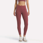A woman stands facing forward in Reebok Women's Odyssey High-Rise 7/8 Leggings in Blackberry, paired with a matching sports bra and white athletic shoes, arms relaxed at her sides against a plain white background.
