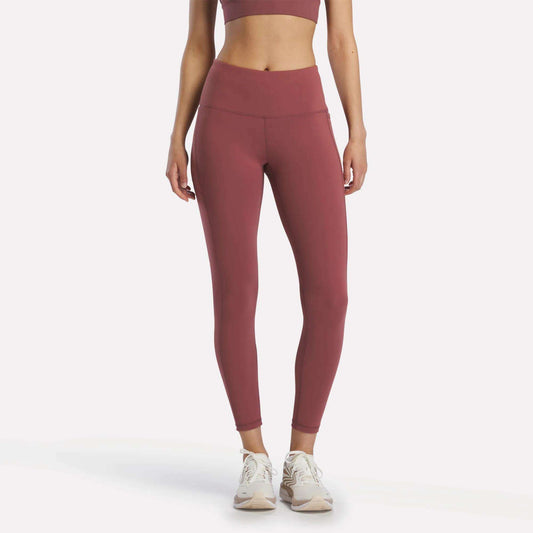 A woman stands facing forward in Reebok Women's Odyssey High-Rise 7/8 Leggings in Blackberry, paired with a matching sports bra and white athletic shoes, arms relaxed at her sides against a plain white background.