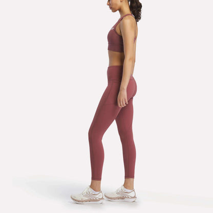A woman in profile faces left, wearing Reebok Women's Odyssey High-Rise 7/8 Leggings and a matching sports bra, paired with white athletic shoes, set against a plain white background.