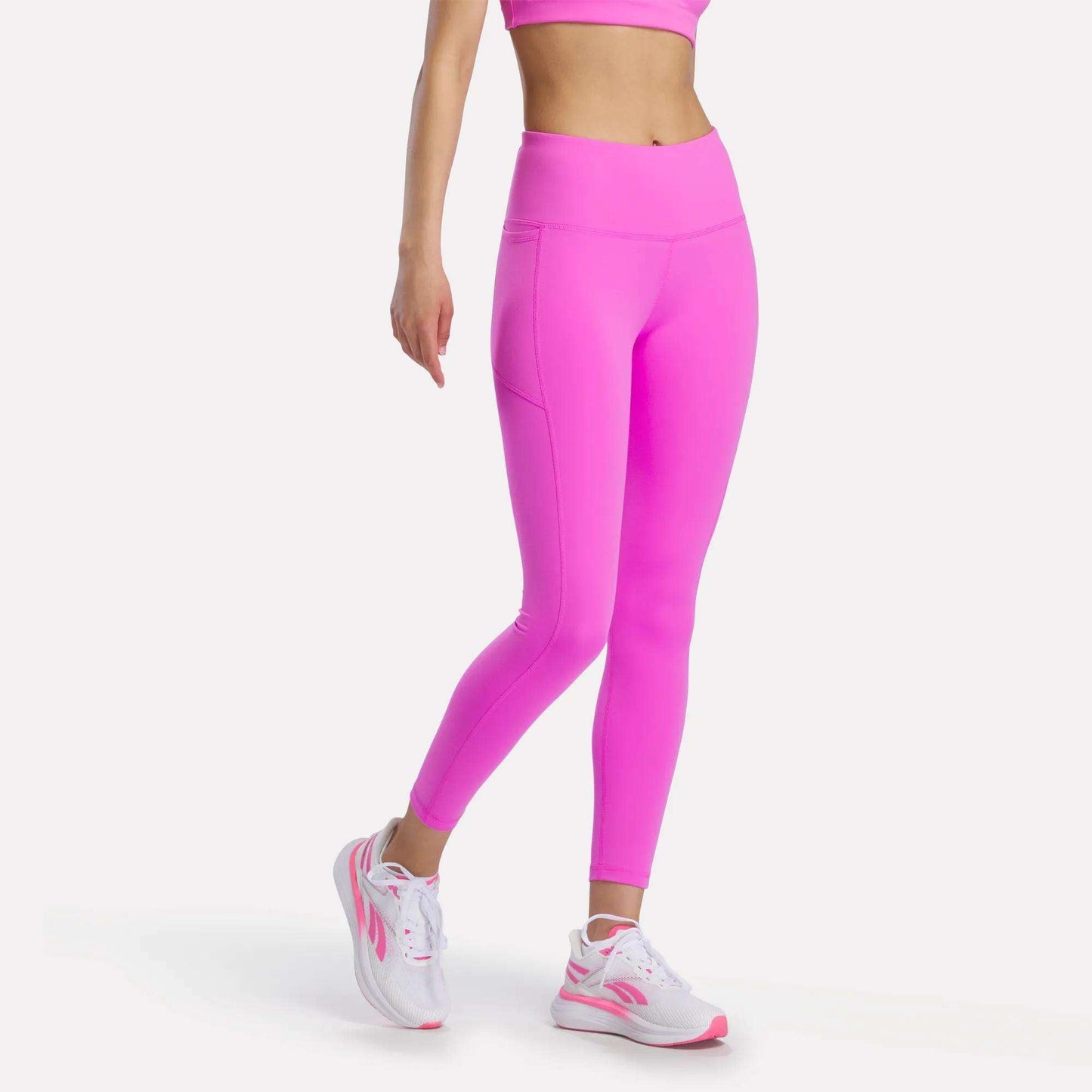 A person wears Reebok Women's Odyssey High-Rise 7/8 Leggings in Vector Virtual Pink, paired with a bright pink sports bra and white-pink athletic shoes, standing against a plain white background with their head and right hand out of frame.
