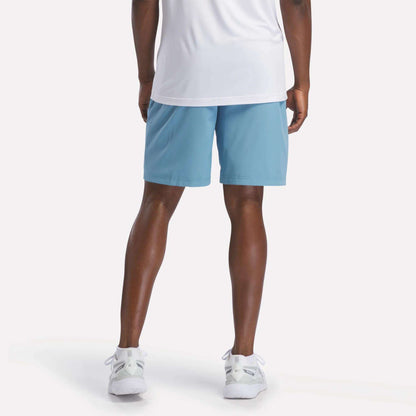A person wearing a white shirt, Men's Range Shorts 9" from Reebok in light blue, and white sneakers stands facing away from the camera, displaying the back of their outfit against a plain white background.