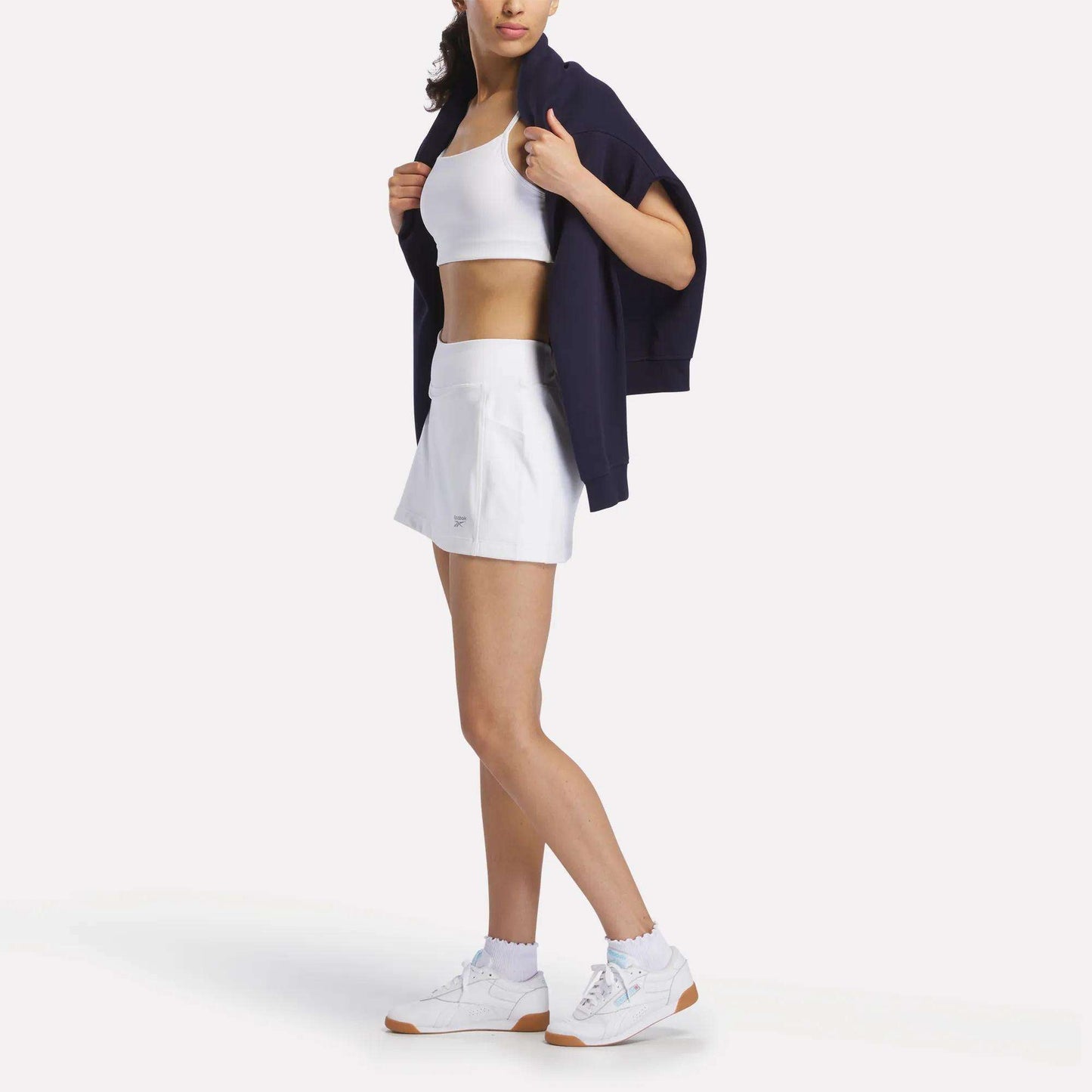 A woman wears a sporty look with a white crop top, Reebok Women's Break Point Skort, white sneakers, and a dark jacket over her shoulders. She stands with her head slightly turned, face partially out of frame.