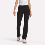 A person stands against a white background, showcasing Reebok Women’s Highstreet High-Rise Pants in black, paired with a white shirt and white sneakers. The image is cropped at the shoulders to highlight the pants.