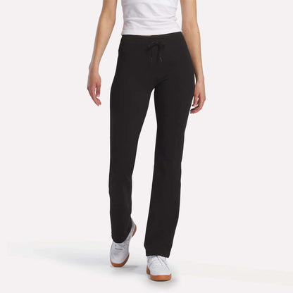 A person stands against a white background, showcasing Reebok Women’s Highstreet High-Rise Pants in black, paired with a white shirt and white sneakers. The image is cropped at the shoulders to highlight the pants.