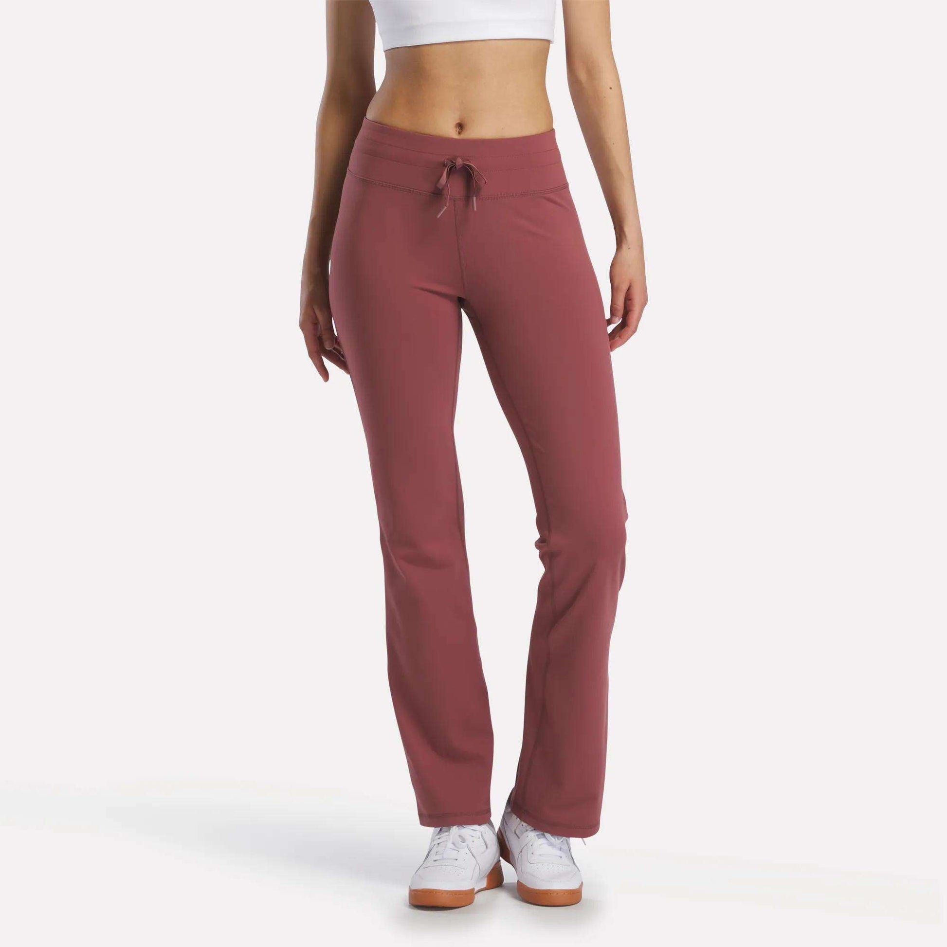 A woman stands against a plain white background, wearing Reebok Women's Highstreet High-Rise Pants in mauve, a white crop top, and white sneakers. Her face is not visible.