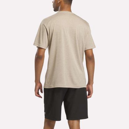A man stands facing away, wearing the Reebok Men's Director Short Sleeve Top in beige with stretchy short sleeves and black shorts against a plain white background.