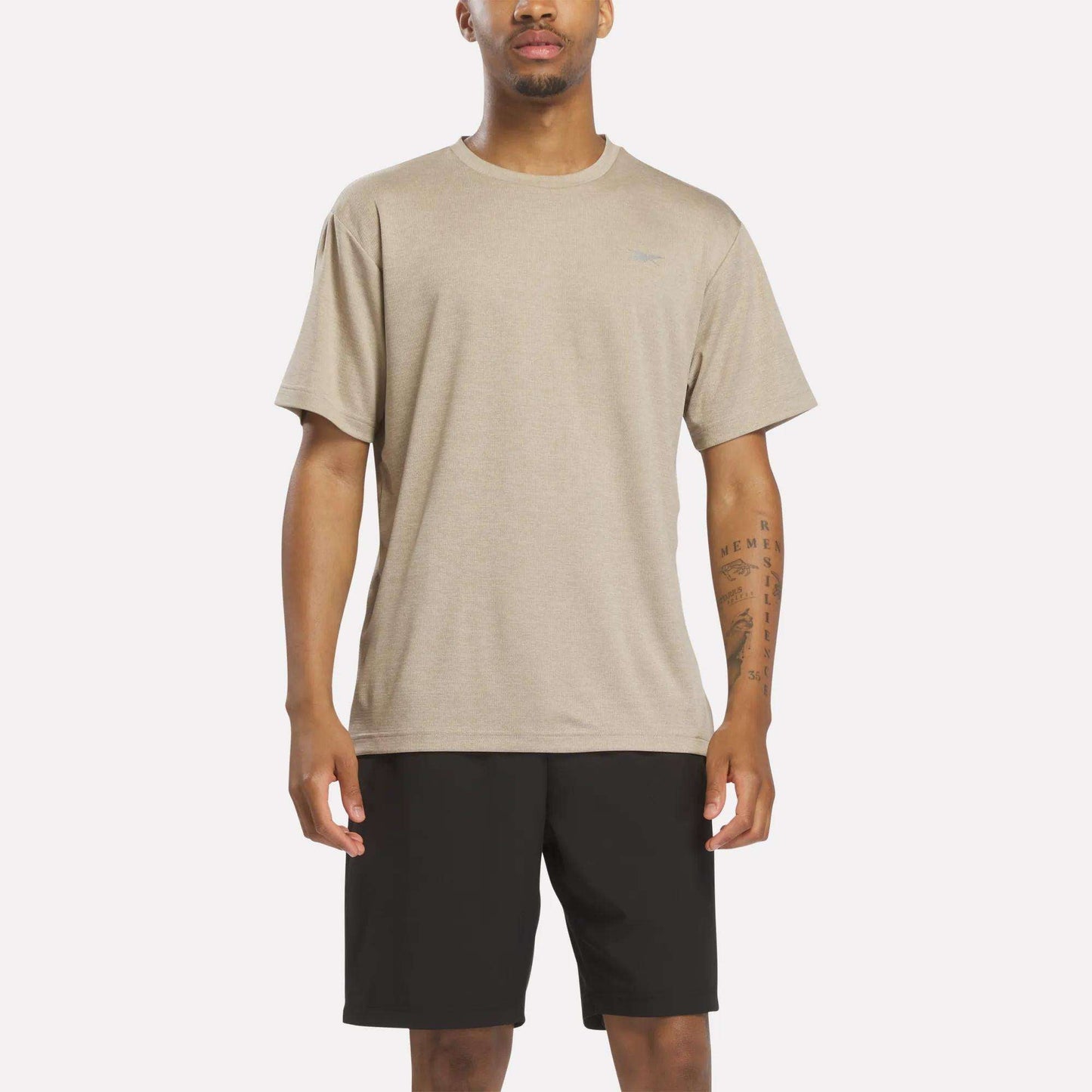 A man stands facing forward wearing a Reebok Men's Director Short Sleeve Top in Grainy Grey Heather and black shorts against a plain white background.