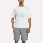 A person wearing a white Reebok X F45 Training Graphic T-Shirt—an ideal men's workout shirt by Reebok—and gray shorts stands against a plain background, with one hand in their pocket.