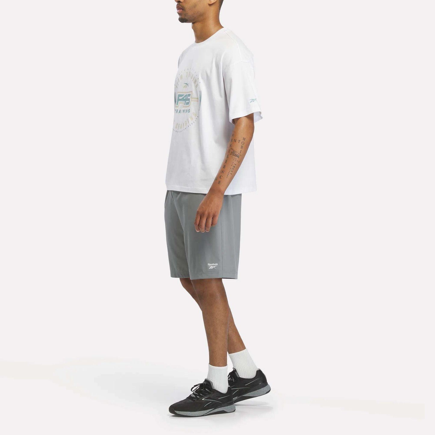 A person stands sideways against a plain white background, wearing a white Reebok X F45 Training Graphic T-Shirt, gray athletic shorts, white socks, and black sneakers.