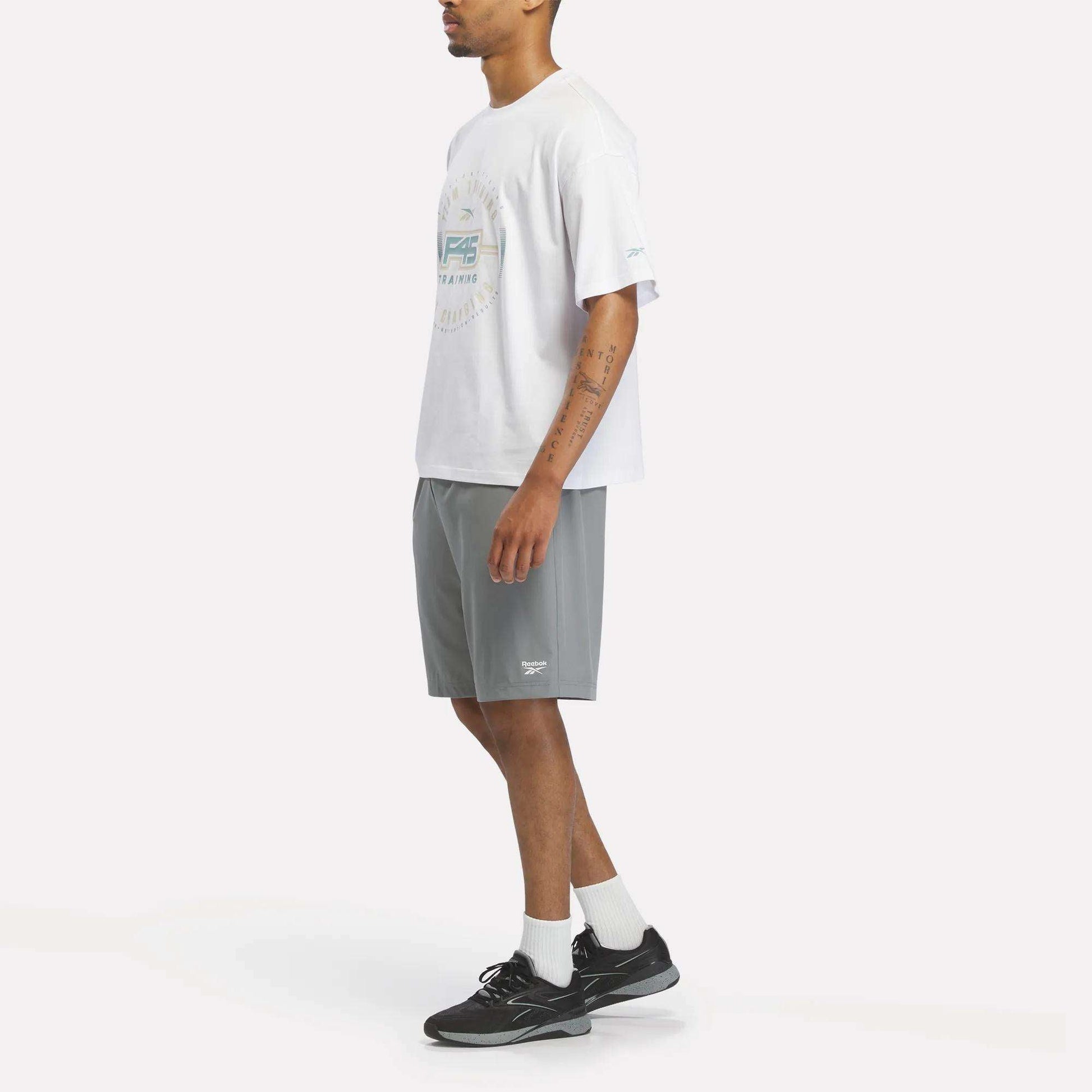 A person stands sideways against a plain white background, wearing a white Reebok X F45 Training Graphic T-Shirt, gray athletic shorts, white socks, and black sneakers.