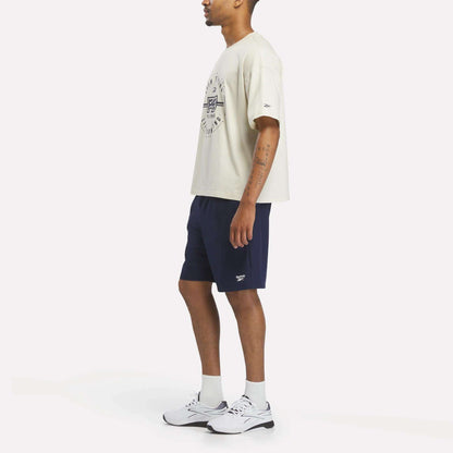A person stands sideways wearing a Reebok X F45 Training Graphic T-Shirt in beige, paired with navy shorts, white socks, and white sneakers. The background is plain white.