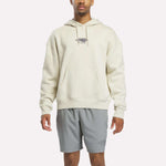 A person wearing the Reebok x F45 Training Relaxed Fleece Hoodie in Linen—a cream-colored, relaxed-fit hoodie featuring "F45 Training" on the chest—paired with light gray athletic shorts, stands against a plain white background.