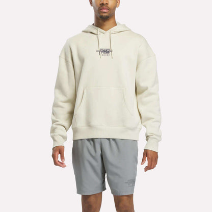 A person wearing the Reebok x F45 Training Relaxed Fleece Hoodie in Linen—a cream-colored, relaxed-fit hoodie featuring "F45 Training" on the chest—paired with light gray athletic shorts, stands against a plain white background.