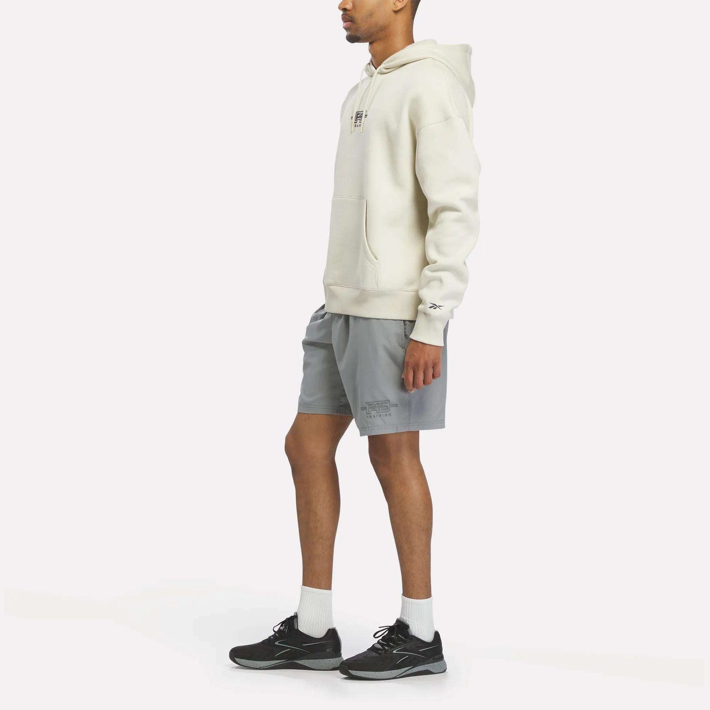 Wearing a Reebok x F45 Training Relaxed Fleece Hoodie in cream, gray shorts, white socks, and black sneakers, a person stands facing sideways against a plain white background.