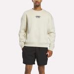 A person stands against a plain white background wearing the Reebok x F45 Training Relaxed Graphic Sweatshirt in Linen with a small chest logo, paired with black shorts.