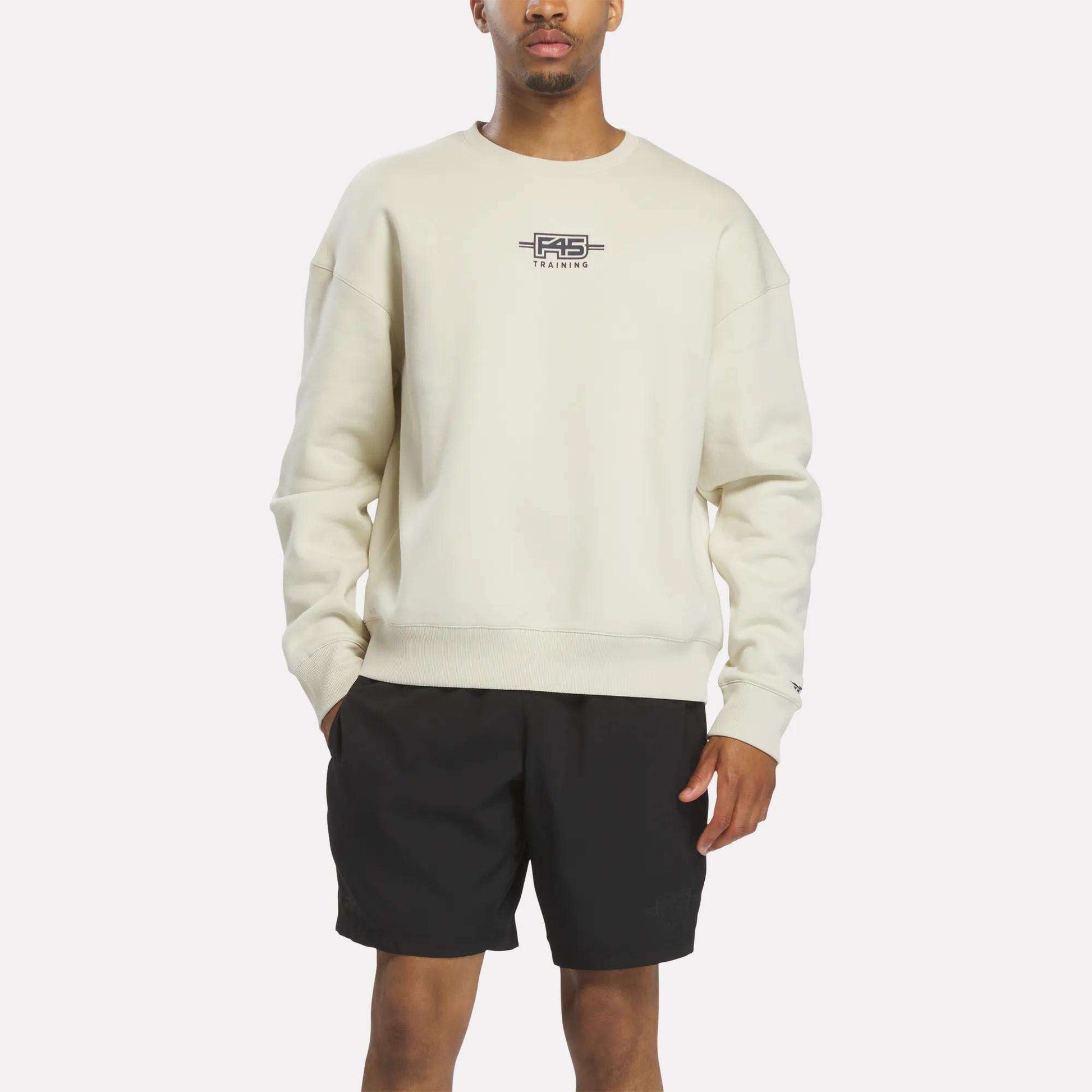A person stands against a plain white background wearing the Reebok x F45 Training Relaxed Graphic Sweatshirt in Linen with a small chest logo, paired with black shorts.