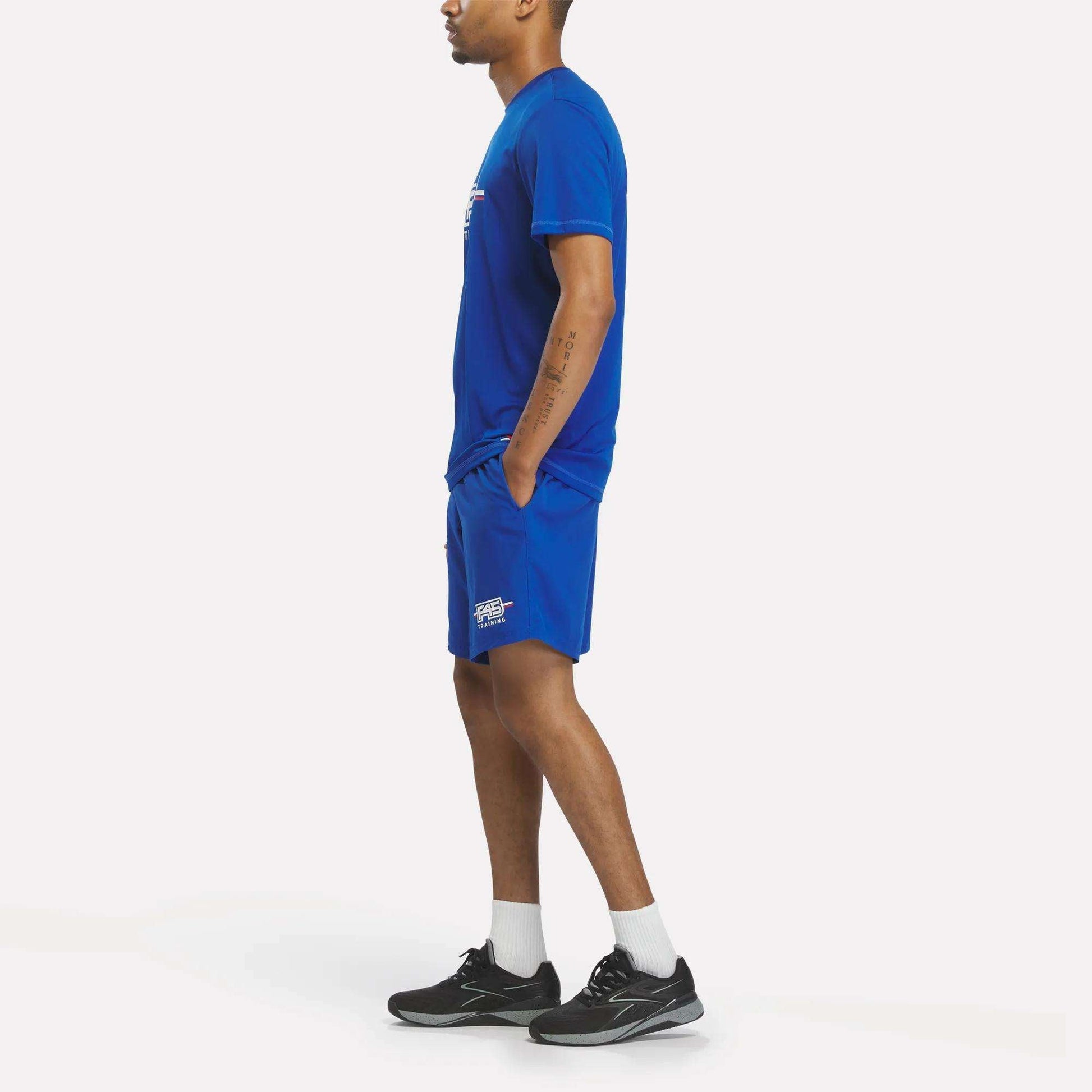 A man stands sideways wearing a Men's Reebok x F45 Training T-Shirt in blue, paired with matching blue shorts, white socks, and black athletic shoes against a plain white background.