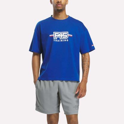 A person wearing a Reebok x F45 Training Graphic T-Shirt in Vector Blue and gray shorts stands facing forward with one hand in their pocket against a white background.