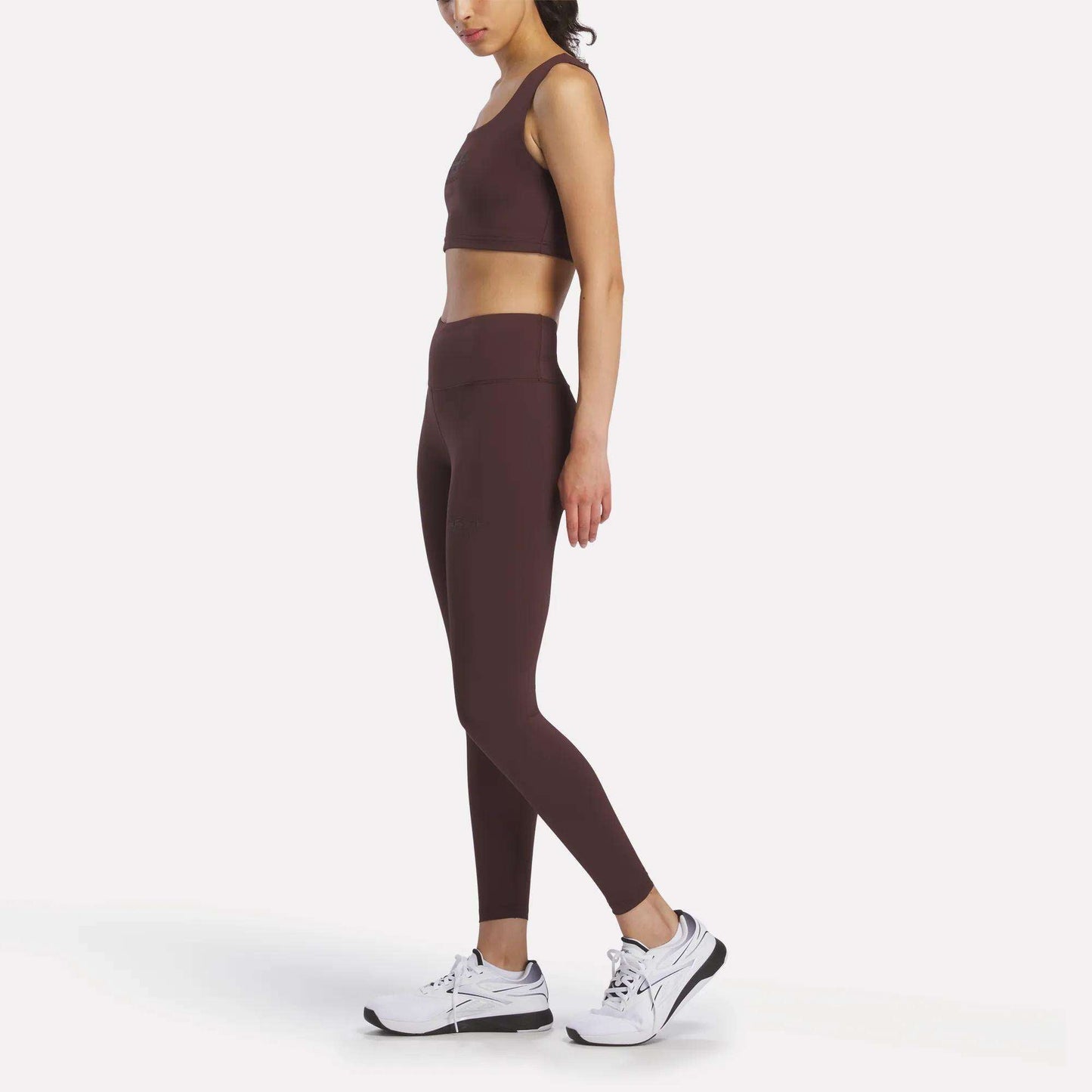 A woman stands sideways against a plain white background, wearing brown athletic leggings and the Women's Reebok x F45 Training Sports Bra by Reebok, paired with white sneakers.