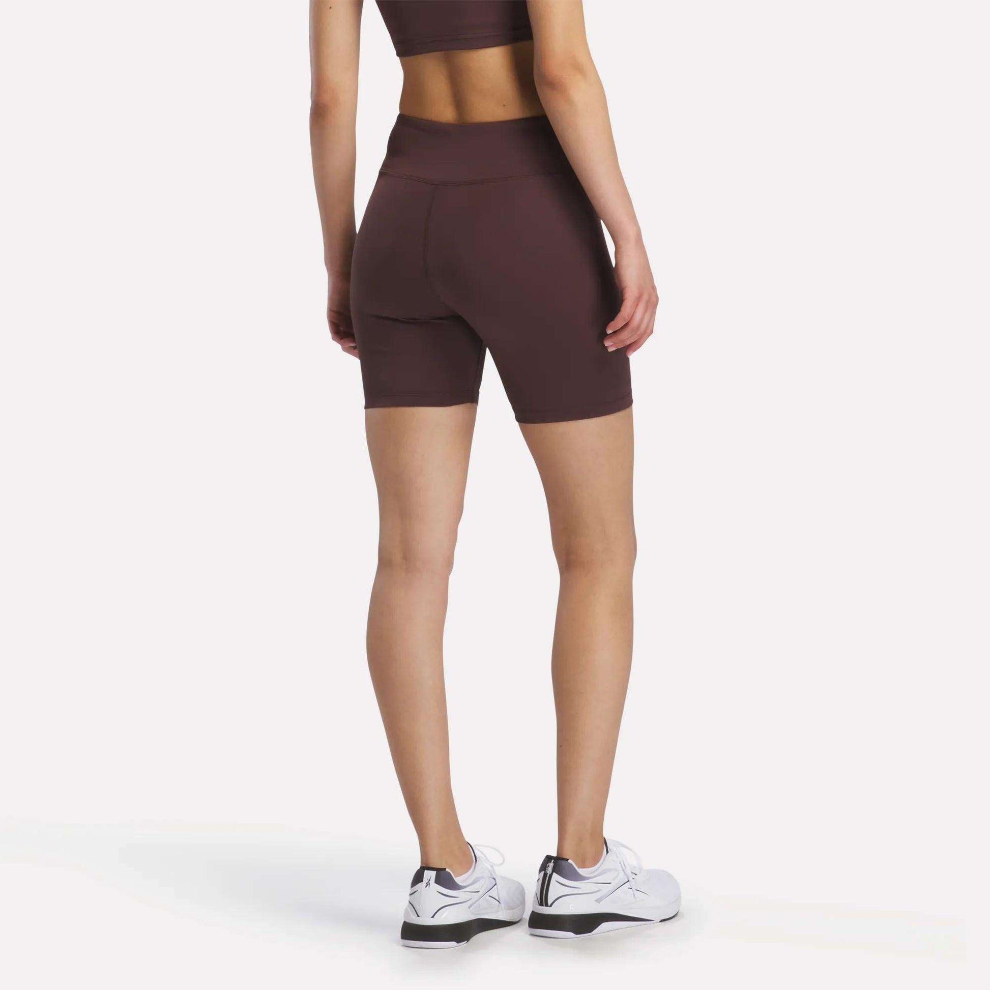 A person stands facing away, displaying the back view while wearing the Women's Reebok x F45 Training High-Rise Biker Shorts in burgundy with a matching sports bra. They are also wearing white Reebok athletic shoes, and the background is plain white.
