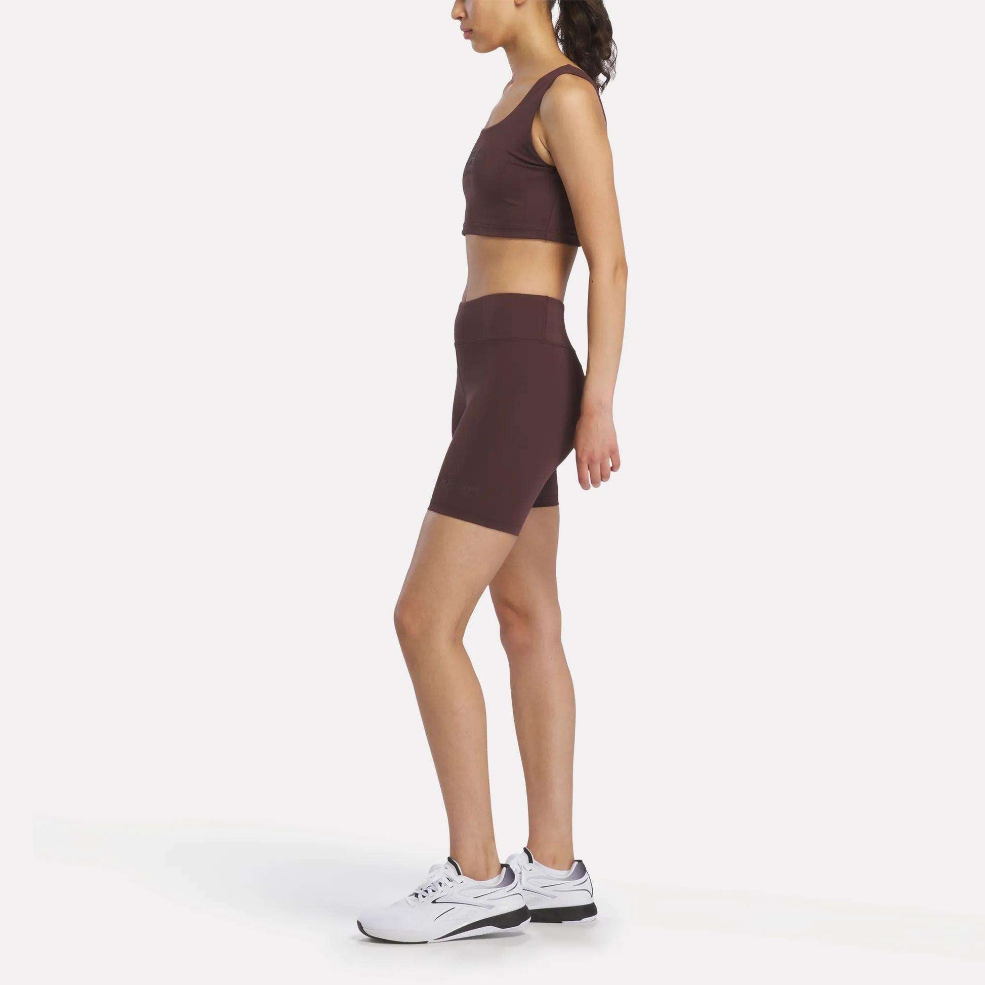 A woman stands in profile against a plain white background, wearing maroon Women's Reebok x F45 Training High-Rise Biker Shorts and a matching sports bra with white athletic shoes, ready for her next F45 Training session.