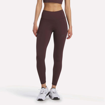 Wearing the Women’s Reebok x F45 Training Leggings and a maroon sports bra, a woman stands with her arms relaxed, paired with white Reebok performance athletic shoes against a plain background.