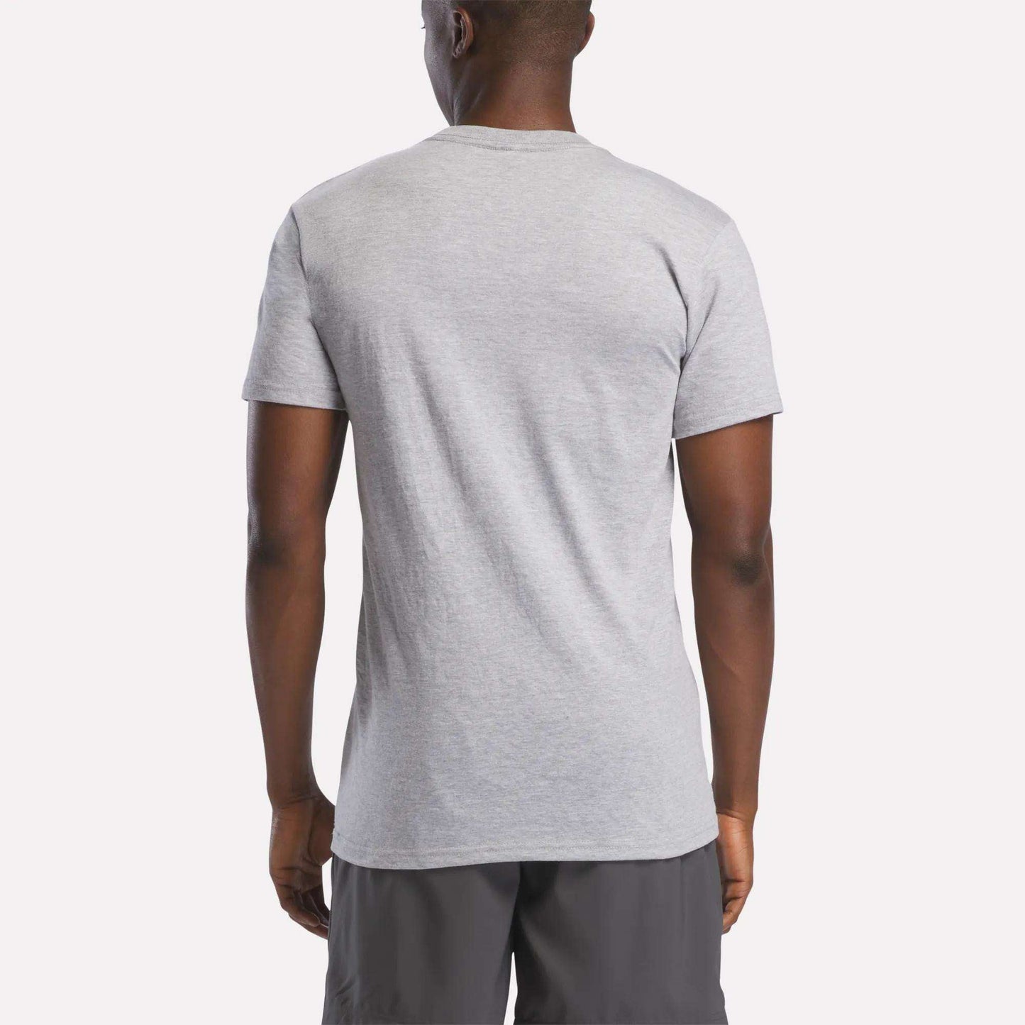 A man, back to the camera, wears a light gray Reebok Men's Stacked Vector Graphic T-Shirt made from lightweight 100% cotton with dark gray shorts, set against a plain light background.