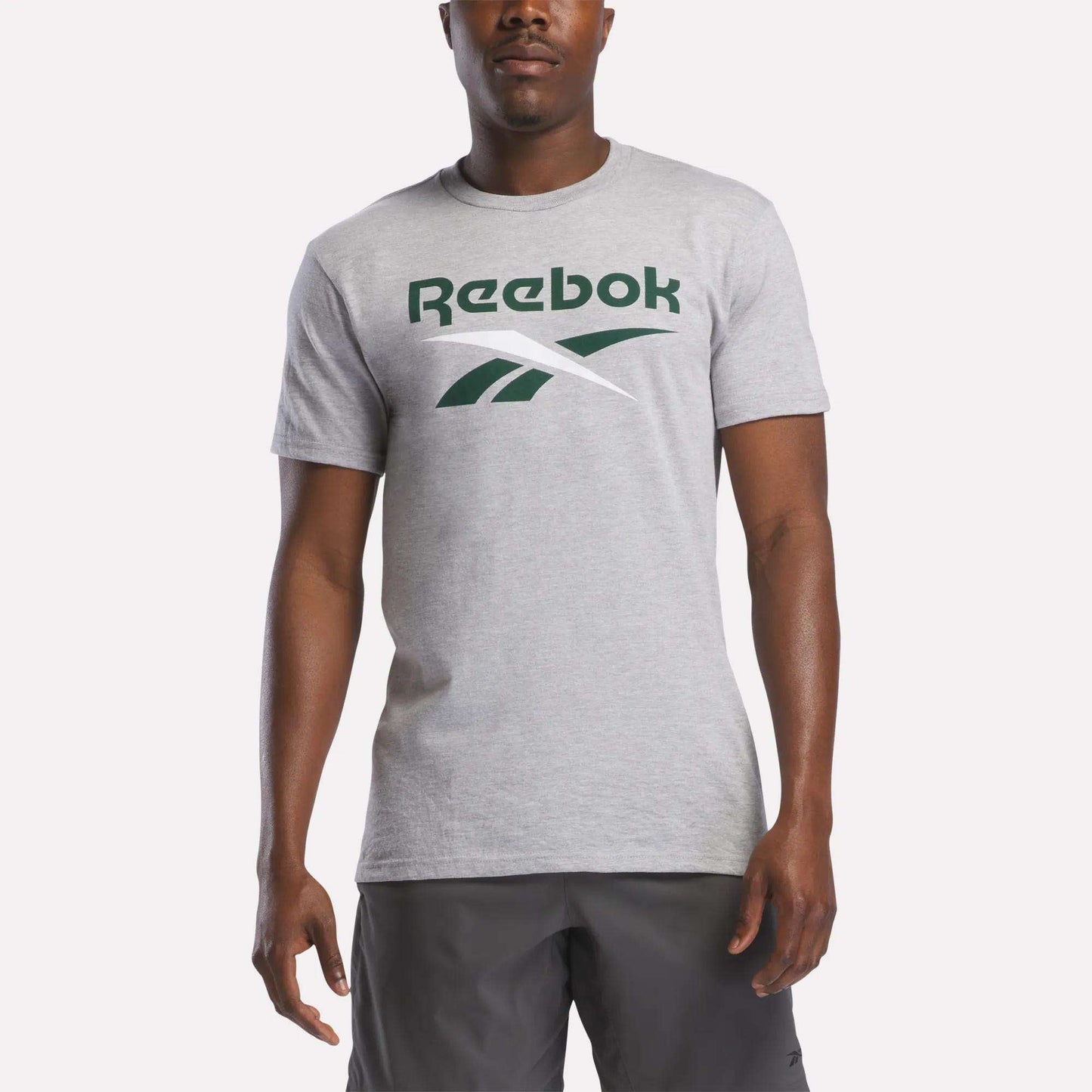 A man wears a Reebok Men's Stacked Vector Graphic T-Shirt in light gray, 100% cotton, with a large green Reebok logo and dark gray shorts, standing against a plain light background.