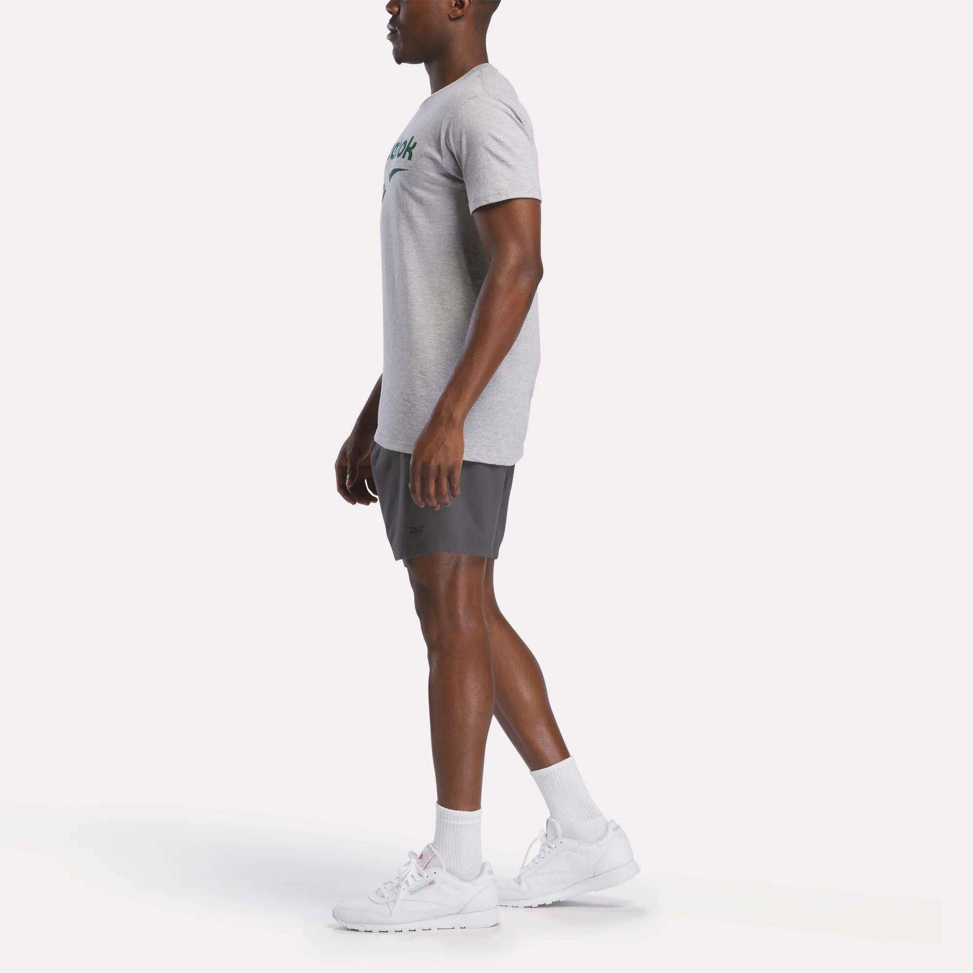 A man stands in profile against a plain background, wearing a gray Reebok Men's Stacked Vector Graphic T-Shirt, dark gray shorts, white socks, and white athletic shoes.